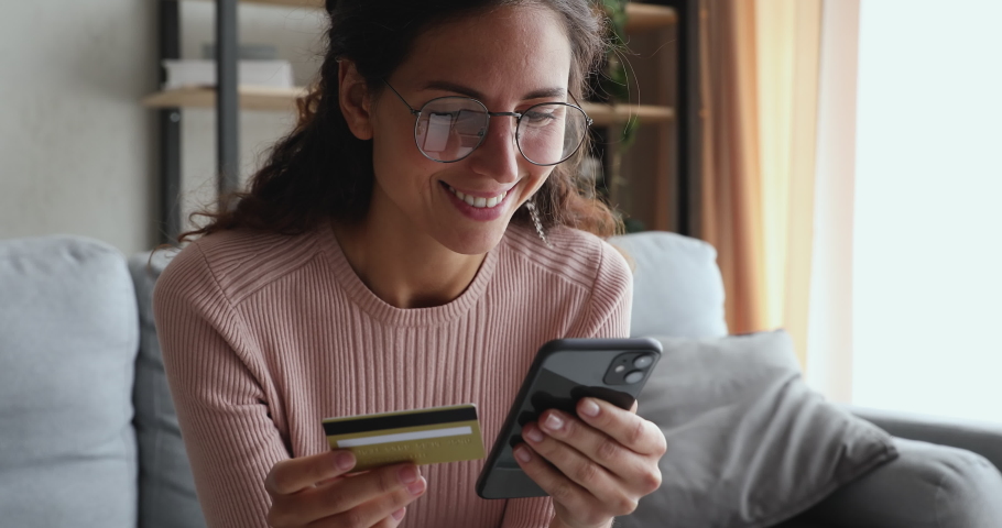 Head shot smiling female client in glasses holding banking credit card involved in verification process in mobile application. Happy young woman doing payments online or shopping in internet store. | Shutterstock HD Video #1055020379