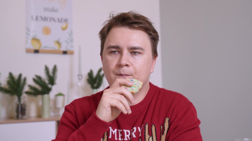 Young man in cozy winter sweater eats cookies in shape of Christmas tree decorated with colored icing, and nods approvingly, enjoying delicious dessert.