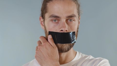 Close up face portrait man looking at camera serious and tearing tape from her mouth on grey background human shut silence face fear expressive attitude taboo censorship violence scared slow motion