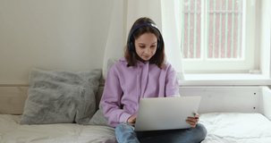 Happy teenage girl sitting on bed in casual clothes wearing headphones using computer, chatting with friends or watching funny video. Young female teen student involved in e-learning by laptop apps.