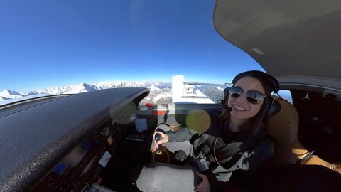 Young female pilot with sunglasses flying a small propeller plane, looking around and smiling. Cockpit view. Aerial shot from high altitude. POV.