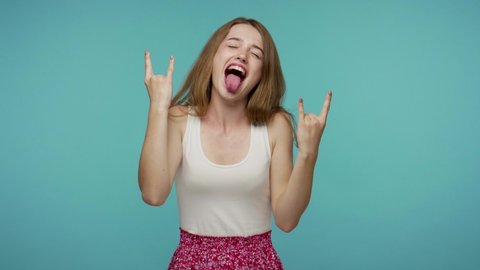 Carefree delighted beautiful girl demonstrating tongue out and rock and roll hand gesture, punk sign, shouting excited by win, crazy about success. indoor studio shot isolated on blue background