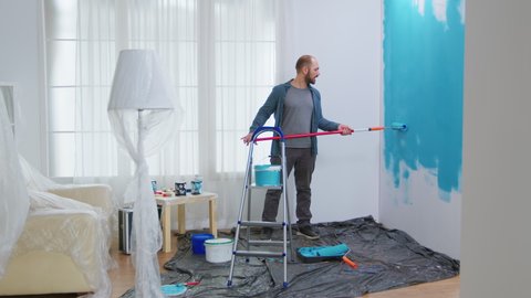 Handyman dancing and painting wall with roller brush while renovating his apartment. Apartment redecoration and home construction while renovating and improving. Repair and decorating.