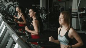 Two happy active girls in sport clothing running on treadmill at fitness center. Beautiful female friends chatting and doing cardio together. Concept of endurance and motivation.