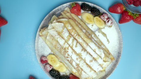 Homemade French Crepes with jam and chocolate cream/sauce in a serving plate with fresh berries: strawberries, blueberries, rose berries, bananas on blue background.