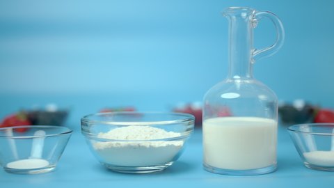 French Crepe ingredients in transparent glass bowls and a jar of milk with berries on background on a blue backdrop. Concept of preparing french crepes/pancakes. Eggs, flour, oil, sugar, milk, salt.