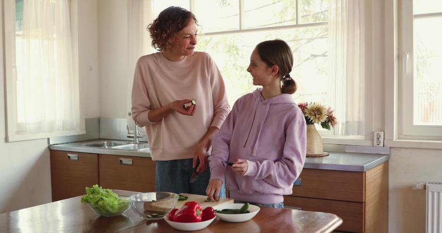 Happy mother teaching teenage child daughter cooking vegetarian food in modern kitchen. Smiling mom controlling how adolescent kid girl preparing food, chopping fresh vegetables on wooden board.