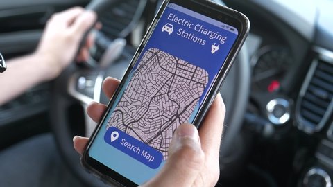 Closeup on a driver's hand holding a smartphone mobile device from inside his vehicle,using an app to search EV charging stations to charge his electric car