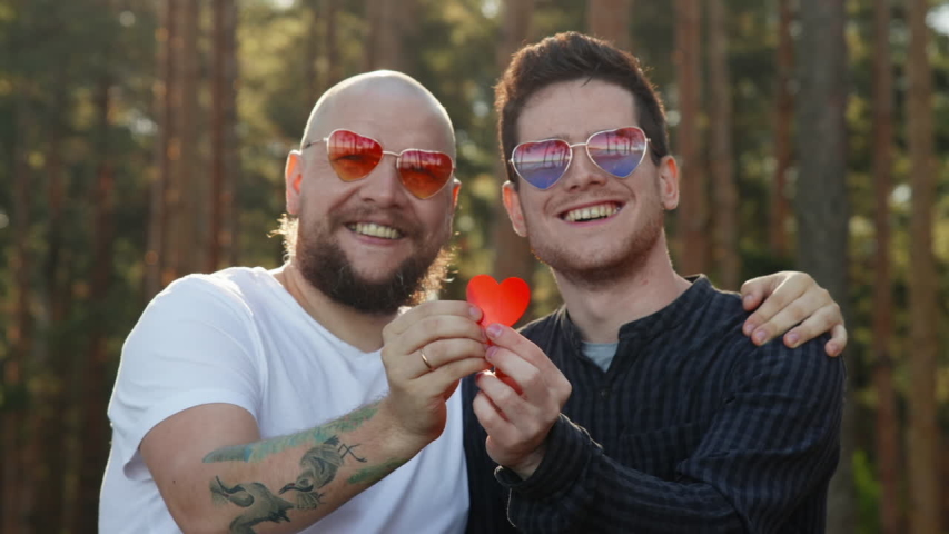 Homosexual couple dating in nature. Portrait of two affectionate gay men in love wearing sunglasses showing paper heart and smiling to camera. LGBTQI, Pride Event, LGBT Pride Month, valentine's day | Shutterstock HD Video #1055069918