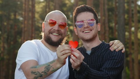 Homosexual couple dating in nature. Portrait of two affectionate gay men in love wearing sunglasses showing paper heart and smiling to camera. LGBTQI, Pride Event, LGBT Pride Month