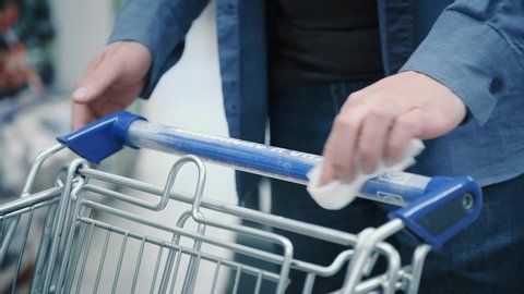 young man in blue shirt cleans handle of shopping cart with wet wipe to remove bacteria in brightly lit supermarket extreme close view