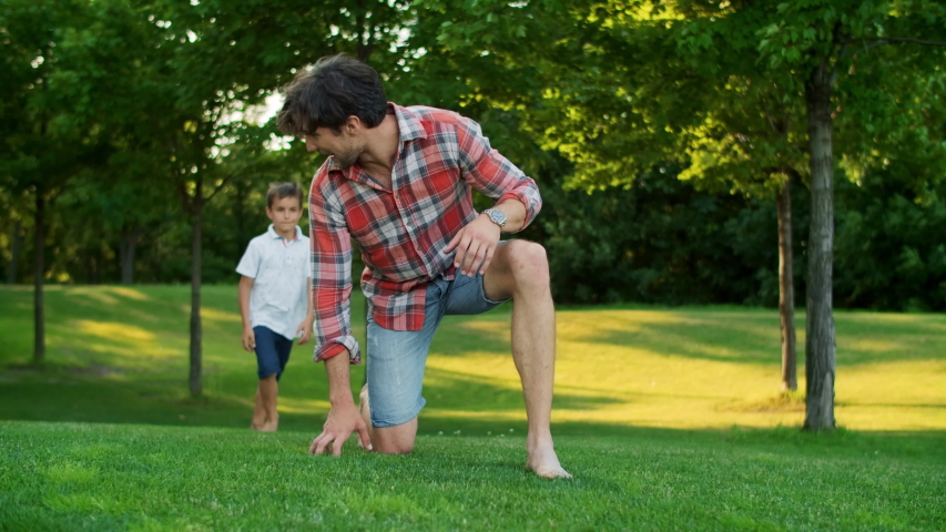 Happy son jumping on father back in green park. Smiling man giving boy piggyback riding outdoors. Cheerful son and father running in field. Positive family spending weekend together outside Royalty-Free Stock Footage #1055074772