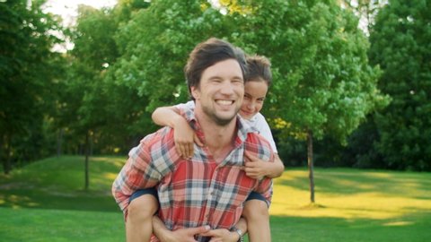 Happy son jumping on father back in green park. Smiling man giving boy piggyback riding outdoors. Cheerful son and father running in field. Positive family spending weekend together outside
