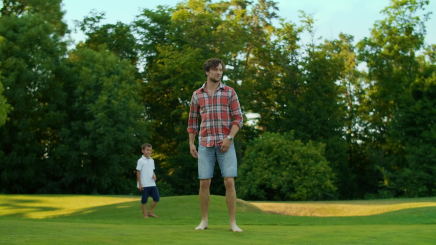 Handsome man making flip in green field in slow motion. Focused guy doing trick in meadow. Young man jumping in air. Happy family standing on summer park background