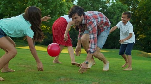 Happy parents and kids playing with ball in green meadow. Smiling man holding ball in hands. Positive family trying to get ball during game. Cheerful family enjoying summer outdoor activity
