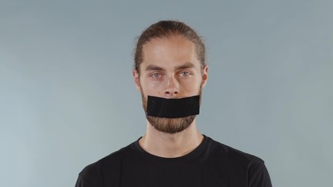 Close up man with black tape on mouth over grey background looking at camera serious human shut silence face fear expressive attitude taboo censorship violence scared slow motion