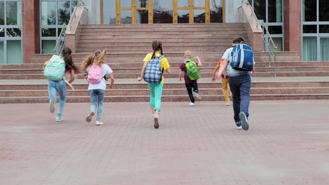 junior schoolchildren with backpacks run to school entrance past flowerbeds backside view slow motion