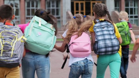 junior students in casual clothes with schoolbags run to school building on warm day backside close view slow motion