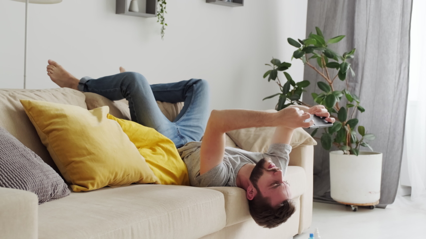 Young bearded man lying on sofa upside down playing mobile game on his smartphone, self quarantine concept Royalty-Free Stock Footage #1055083997