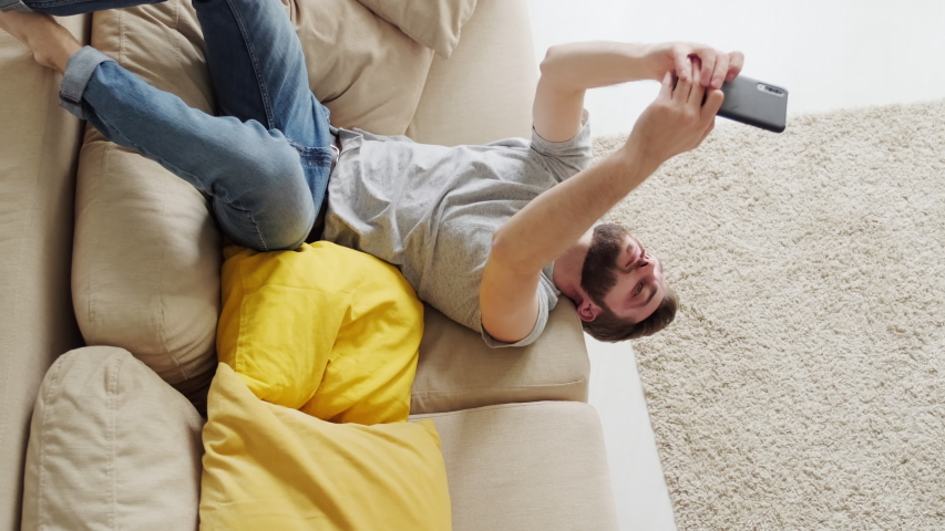 Bearded man lying on sofa upside down playing video game on his smartphone then getting bored and putting his gadget away, lockdown concept Royalty-Free Stock Footage #1055084531