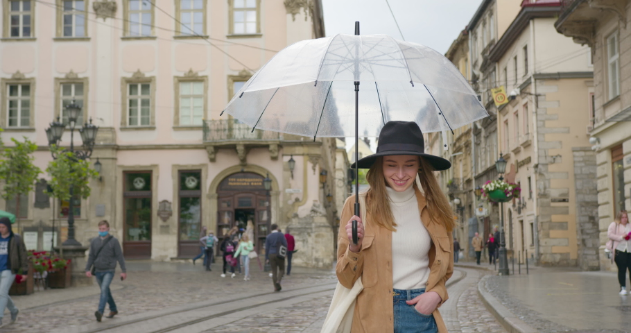A girl is walking slowly in the rain and smiling. She is holding an umbrella in her hand. A little rain is falling. She has a big hat on her head. 4K