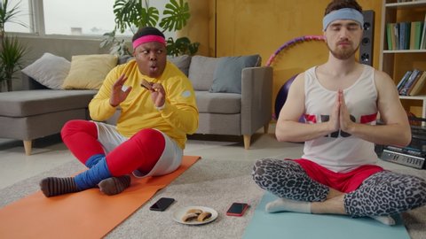 Bearded yoga trainer practicing asana in lotus pose on mat while his student overweight funny african guy eating cookies without permission.