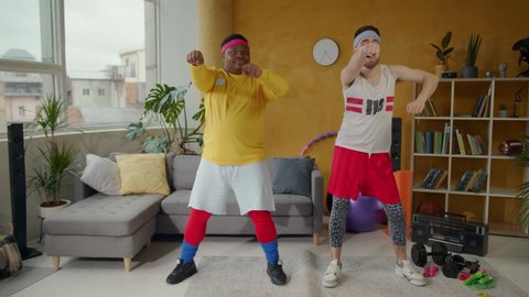 Excited funny retro multi-ethnic couple of unfit friends men wearing sportswear performing warm-up cardio dance exercising indoors.