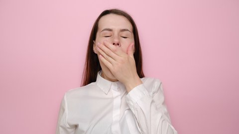 Drowsy young woman yawning, sleepy inattentive, feeling somnolent lazy bored gaping suffering from lack of sleep, dressed in white shirt, isolated on pink background. Tiredness and awakening concept