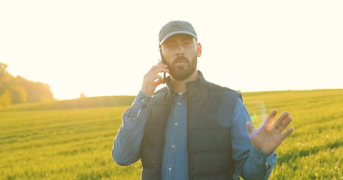 Caucasian young male farmer in hat walking in green field and talking on mobile phone. Attractive man speaking on cellphone in margin in summer and telling something about agricultural business.