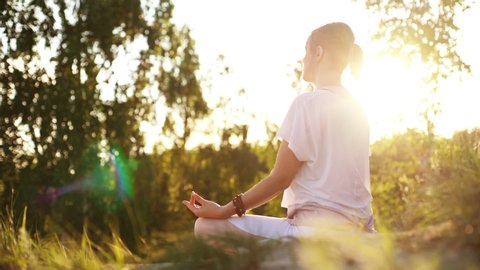 Side view of meditating woman putting hands in namaste mudra posture sitting in lotus pose background of beautiful landscape. Unrecognizable female practicing yoga alone evening sun on nature.