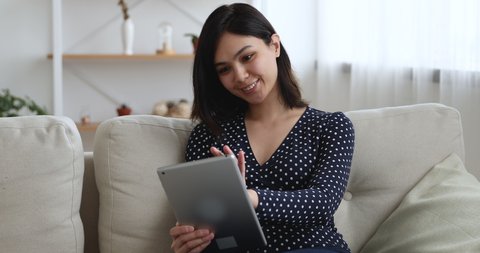 Asian woman sitting on couch in cozy living room hold tablet device smile enjoy free time chat on-line with friends, buy in e-commerce web sites. Modern tech, everyday usage easy and cool app concept