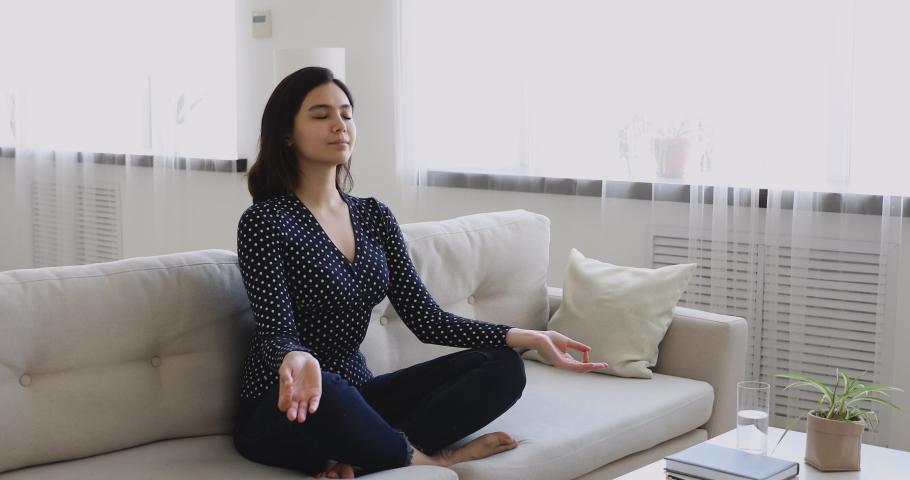 Asian ethnicity millennial woman sit cross-legged on couch with folded fingers, closed eyes do inhale exhale exercise breathing fresh air enjoy mindfulness inner harmony, meditation yoga time concept Royalty-Free Stock Footage #1055094485