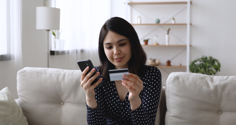 Asian woman sitting on couch holding cellphone using credit card makes instant payment feels happy. Easy on-line shopping, modern tech, safe successful convenient payment, sale and discounts concept Royalty-Free Stock Footage #1055094494
