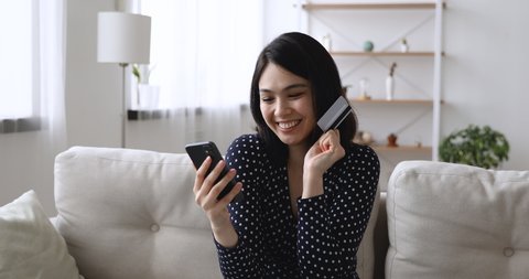 Asian woman sitting on couch holding cellphone using credit card makes instant payment feels happy. Easy on-line shopping, modern tech, safe successful convenient payment, sale and discounts concept