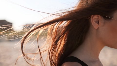 Close back view of brown woman hair tender movement in the air in slow motion 120 fps. Brunette woman in sunglasses walking on sandy beach. Wind blows airy hair shining in sun on sunset. Macro video.
