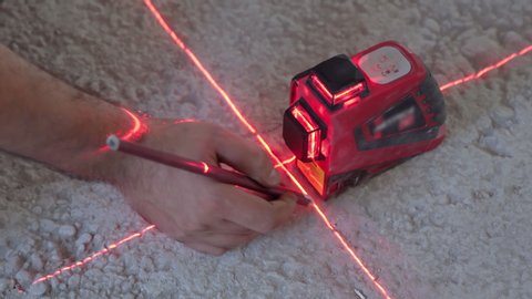 Building worker setting modern laser level on the floor on construction site for further measuring, construction device showing red rays crosshair for taking accurate measurements. Construction