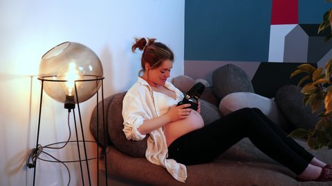 Pregnant woman is sitting and lets her belly listen music in headphones. The expecting mom is sitting at the sofa and moves her leg in the rhythm of music. Theme of pregnancy and life at home.