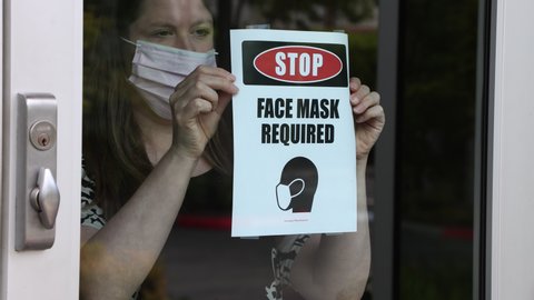 Female shop owner wearing a face mask posts a notice on the glass door entrance requiring face mask for entry