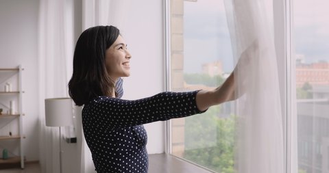 Vietnamese ethnicity young woman come to window in cozy modern living room opens curtains breathes fresh air-conditioned air admires view dreams of the future enjoys morning starting new day concept
