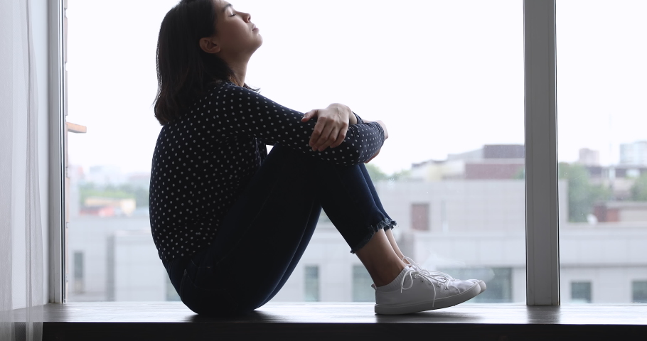 Side view sad Vietnamese woman sitting on windowsill embraces laps feels unhappy, lost on unpleasant thoughts, suffering due life troubles, break up or divorce, waiting or missing beloved man concept | Shutterstock HD Video #1055106491