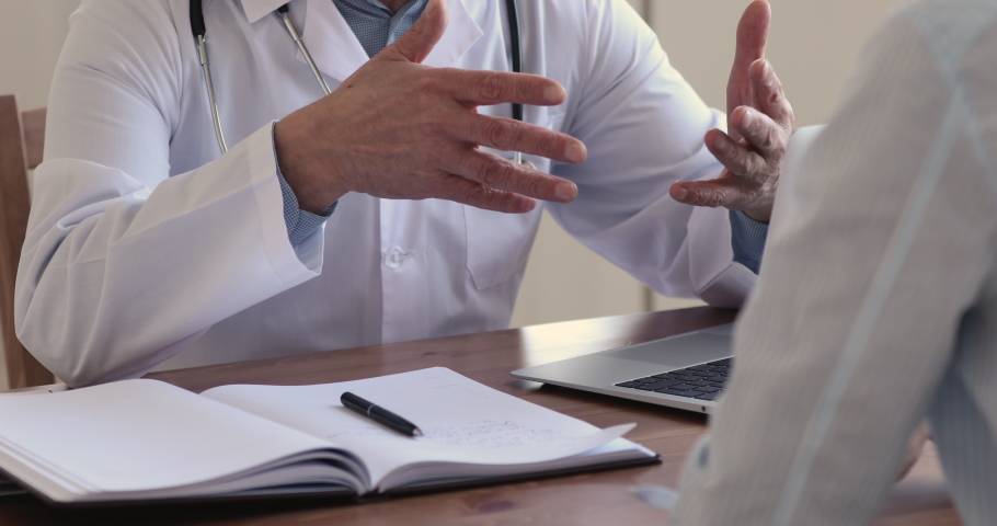 Close up older senior male doctor gesturing, explaining medical treatment to focused patient at checkup meeting in hospital office. Focused middle aged surgeon therapist in coat consulting client. | Shutterstock HD Video #1055106506