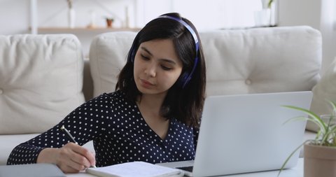 Focused asian woman in headphones using pc sit at coffee table at home, pretty student girl writes notes learn language, do task, watch on-line webinar, listen audio course, e-learn education concept