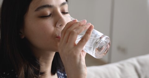 Close up view young vietnamese woman sitting on sofa at home reduces relieve thirst takes a sips drinking whole glass of water. Dehydration prevention, skin and health care, healthy life habit concept