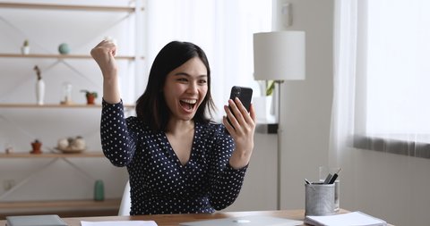 Asian woman sit at desk hold cellphone typing message receive sms from bank feels incredible happy. Girl makes yes I did it gesture look very excited. Achievement, got hired, lottery money win concept