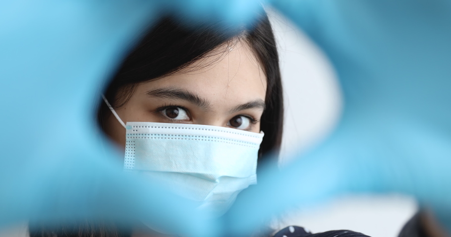 Close up Asian woman in blue medical face mask wears protective gloves bring nearer hands to camera makes heart shape looks through fingers. Take care of yourself at COVID-19 pandemic disease concept Royalty-Free Stock Footage #1055106740