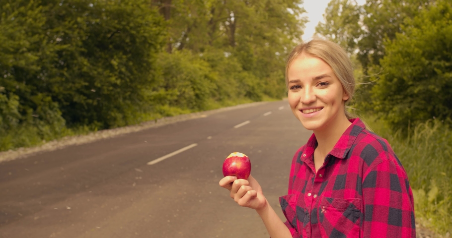 portrait of a hitchhiker woman eating red apple while sitting on backpack outdoor near road