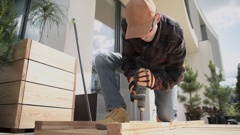 Skilled Male Carpenter Drilling Holes With Cordless Drill In Lumber For Outdoor Residential Project.