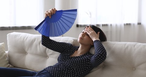 Unhealthy young Vietnamese woman sit leaned on couch at home waves blue waver fan reduce heat feels unwell. Sick millennial girl rest on sofa, suffering from sun stroke or hormonal imbalance concept