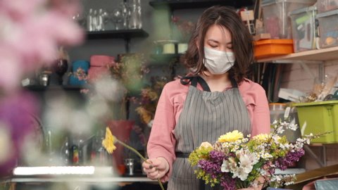 female adding new flower to bouquet. asian woman florist holding a half made arrangement and adding plants to composition. wearing mask due to flu. Designing, floral workshop, working concept