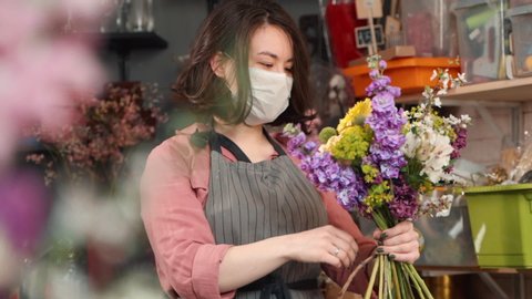 woman in facial protective mask finishing completing bouquet. asian female florist arranging wildflowers in a flower shop or workshop. concept virus prevention, covid19 epidemic